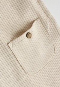 Tela beige acanalada con un bolsillo cosido que presenta un pequeño botón redondo grabado con la palabra "NEXT".