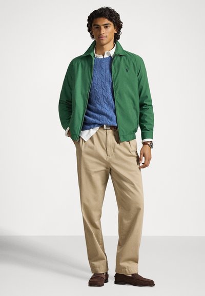 Πράσινο μπουφάν τύπου bomber πάνω από μπλε πουλόβερ με ανάγλυφο πλέξιμο, λευκό πουκάμισο, μπεζ παντελόνι και καφέ παπούτσια από σουέτ. Το μοντέλο στέκεται μπροστά σε φωτεινό φόντο.