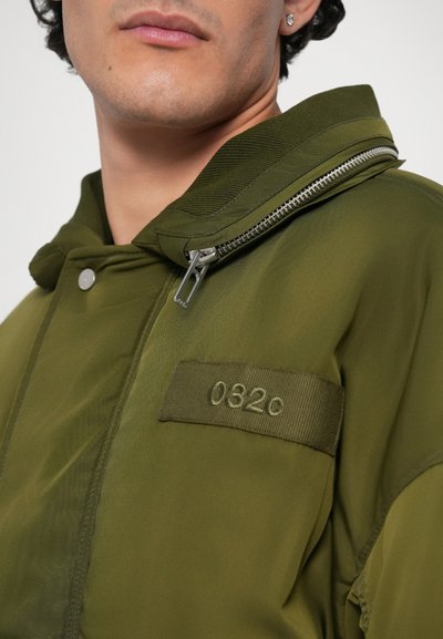 Olijfgroen gewatteerde jacket met capuchon, gemaakt van lichtgewicht materiaal. Beschikt over een ritssluiting, een patch met "0320" en een gladde textuur.