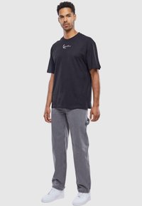 Camiseta de algodón negra con cuello redondo y mangas cortas, combinada con pantalones de mezclilla grises y zapatillas blancas, de pie contra un fondo neutro.