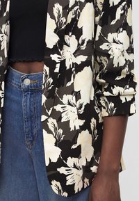 Blazer floral noir et blanc sur un top court noir et un jean bleu taille haute, bras détendu sur le côté.