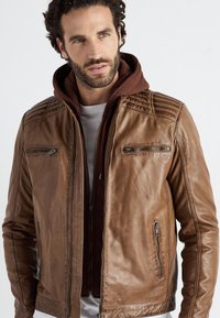 Ruskea nahkatakki, jossa on urheilulliset olkapäät, vetoketjulliset taskut ja huppukasvi sisältä. Sileä pinta ja istuva muotoilu korostavat sen modernia ilmettä.