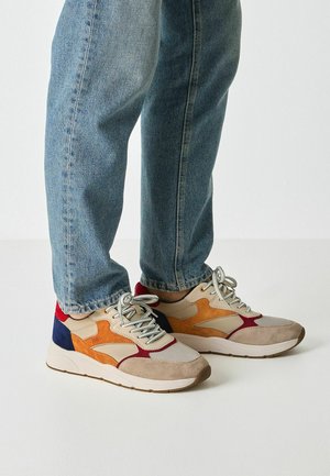 Šarene tenisice s bež antilopom, narančastim i tamnoplavim detaljima, teksturiranim mrežastim materijalom i kremastim gumenim đonom, u kombinaciji s plavim trapericama.