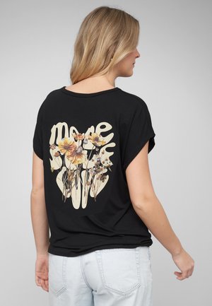 Lyshåret kvinde iført en sort t-shirt med blomstergrafik og beige abstrakt tekst på ryggen, kombineret med lyseblå jeans.