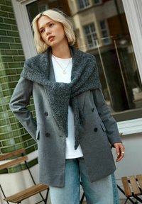 Gray double-breasted coat with a textured dark gray scarf draped over it. White shirt underneath, paired with light blue jeans. Minimalist design.