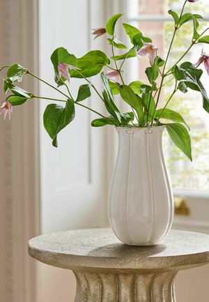 Hvid keramisk vase med grønne løvgrene og små lyserøde blomster på en struktureret rund stensokkel foran et vindue.