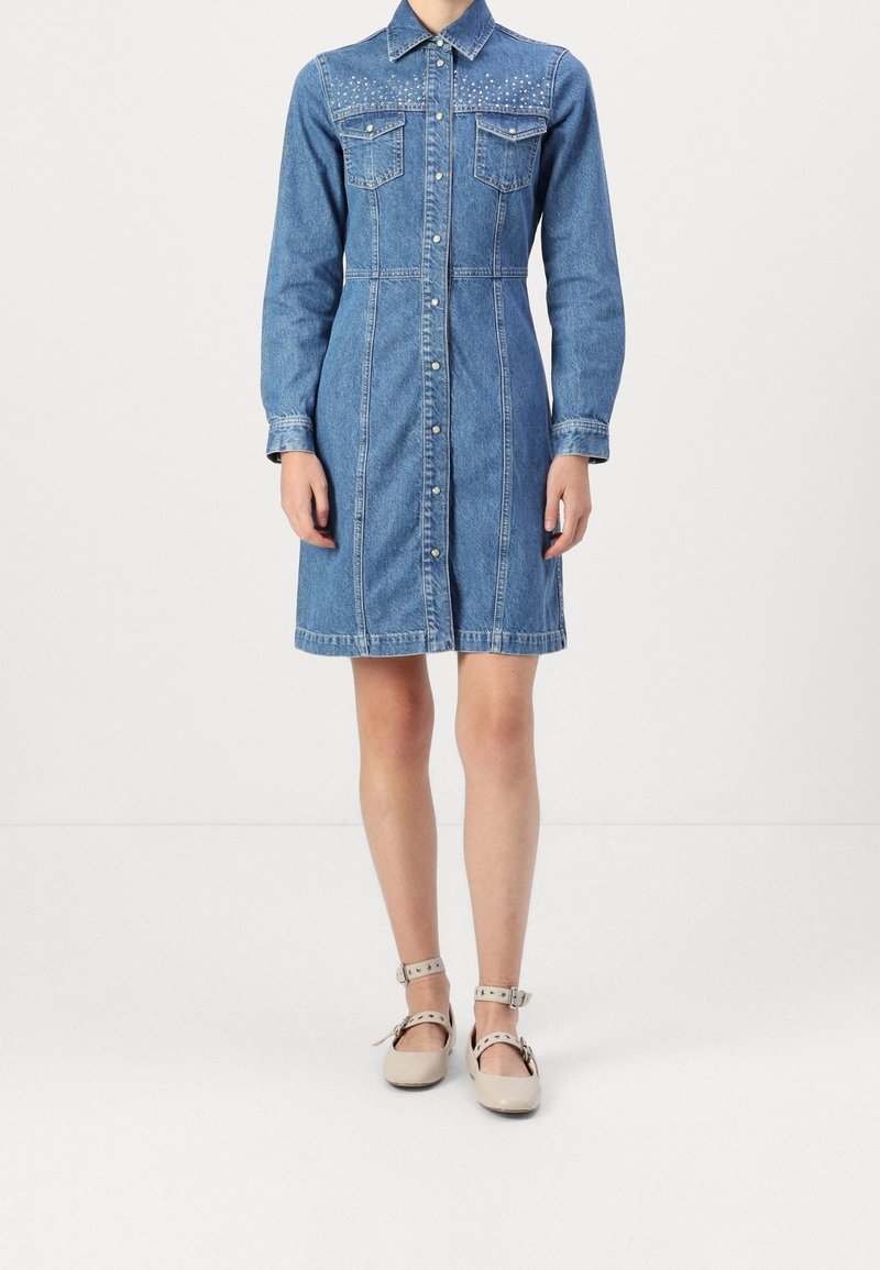 Donna che indossa un vestito di jeans blu al ginocchio con maniche lunghe, chiusura frontale con bottoni e spalline borchiate, abbinato a scarpe beige con cinturino alla caviglia.