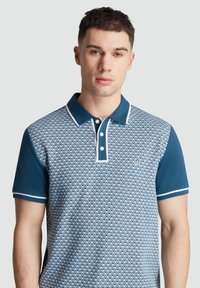 Polo bleu avec un motif en losanges, manches unies contrastantes, piping blanc au col et un petit logo sur la poitrine.
