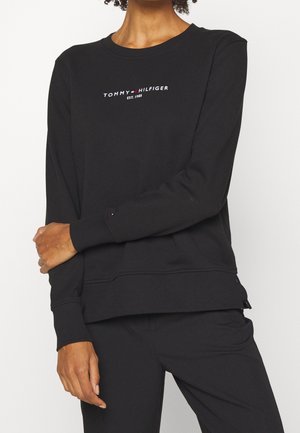 Personne portant un sweat-shirt noir à manches longues avec le logo "Tommy Hilfiger Est. 1985" sur la poitrine et un pantalon noir assorti.