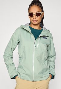 Svetlo zelena vremensko odporna jakna s kapuco, žepki z zadrgo in sivimi poudarki. Gladek teksturni in zračen dizajn tkanine.