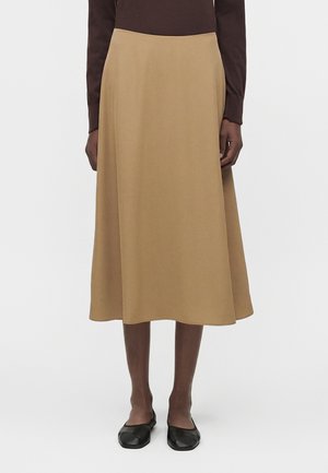 Άτομο που φοράει μπεζ midi φούστα σε γραμμή Α, καφέ μπλούζα με μακριά μανίκια και μαύρα επίπεδα παπούτσια, στέκεται μπροστά σε απλό φωτεινό φόντο.