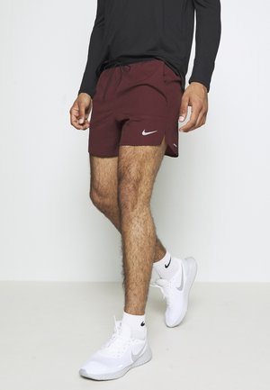 Hombre con pantalones cortos deportivos Nike color granate, camiseta de manga larga negra, calcetas blancas Nike y zapatillas de correr blancas Nike sobre fondo liso.