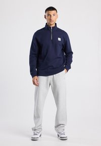 Navy zip pullover with a stand collar, featuring a small logo patch, paired with light gray sweatpants and white sneakers.