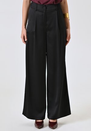 Pantaloni neri a gamba larga in tessuto satinato, con vita alta, pieghe frontali e chiusura a bottone, abbinati a scarpe color borgogna.