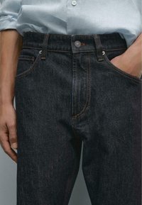 Nærbillede af en person iført mørke denimjeans med orange syninger, hænder delvist i lommerne, mod en neutral baggrund.