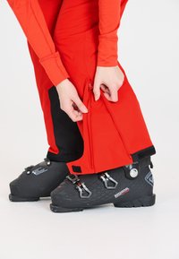 Rode skibroek met een ritssluiting bij de enkel, gecombineerd met zwarte skibotten met verstelbare gespen en een gestructureerd oppervlak.