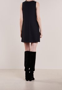 Musta hihaton mekko, jossa on suora leikkaus ja takana vetoketju, yhdistetty polvipitkiin mustiin mokkanahkasaappaisiin, joissa on paksut korot.