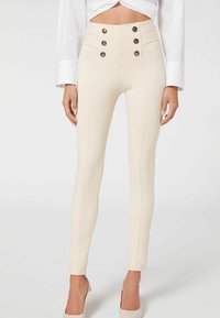 Gros plan sur une femme portant un pantalon beige taille haute avec six boutons décoratifs et des jambes fuselées, associé à une chemise blanche à manches longues et des talons beiges.