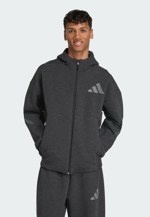 Haut de survêtement de sport gris foncé avec fermeture éclair sur le devant, capuche et coupe décontractée. Présente de grands logos en gris clair sur la poitrine et les manches.