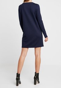 Kvinna klädd i en långärmad marinblå minikjol och svarta högklackade ankelboots, stående med ryggen mot kameran på en vit bakgrund.
