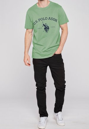 Grøn bomulds-T-shirt med U.S. Polo Assn. logo i marineblå, rund halsudskæring, korte ærmer, kombineret med sorte slim-fit jeans og hvide sneakers.