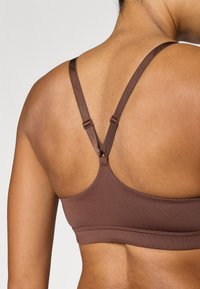 Καφέ αθλητικό σουτιέν με ρυθμιζόμενους ιμάντες και σχέδιο λογοτύπου. Ομαλή υφή και μινιμαλιστικός σχεδιασμός, εστιασμένος στην άνεση και την εφαρμογή.