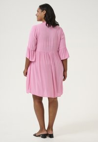 Robe rose, longueur genou, avec des manches volantes, en tissu texturé et coupe ample. Portée avec des sandales à talons blocs noires.