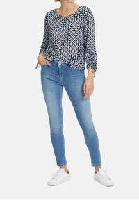 Blouse bleue à motif, coupe loose avec des détails à nouer sur les manches, associée à un jean slim en denim clair et des baskets blanches.