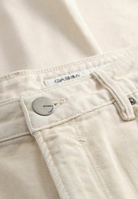 Ανοιχτό μπεζ τζιν υφασμένο από denim υλικό με εμφανές μεταλλικό κουμπί με εγχάρακτο σχέδιο, ραμμένες ραφές και ετικέτα προσαρτημένη στη μέση.