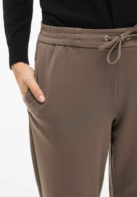Brązowe spodnie z elastycznym pasem i troczkiem, wyposażone w boczne kieszenie i gładką fakturę. Tkanina wydaje się miękka i lekka.