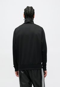 Čierna bunda na zips s vysokým golierom, vyrobená z hladkej látky, s rebrovanými manžetami a spodným lemom, nosená cez zodpovedajúce čierne nohavice.