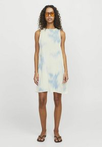 En ärmlös klänning i knälängd i ljusgul med blå batikmönster. Bärs med bruna flip-flop-sandaler och orange solglasögon.