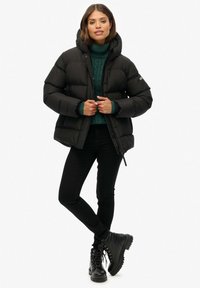 Zwarte gewatteerde jas met een capuchon en ritssluiting, voorzien van een gestikte textuur. Gepaarde met een groene kabelgebreide trui en zwarte skinny jeans.