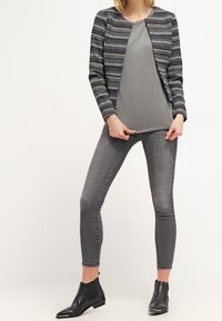 Šedá vzorovaná bunda se strukturovaným designem, nošená přes šedý top, v kombinaci se šedými skinny džíny a černými kotníkovými botami.