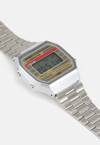 Montre numérique argentée avec un cadran rectangulaire, présentant un design jaune et rouge, un bracelet en acier inoxydable et divers boutons de fonction sur les côtés.