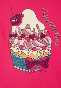 Różowy t-shirt z kolorową grafiką babeczki z błyszczącymi akcentami, wisienką na wierzchu i napisem "Ciesz się małymi rzeczami" w lśniących literach.