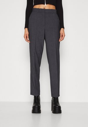 Femme portant un pantalon gris foncé taillé sur mesure avec des plis à l'avant et des bottines noires à plateforme, debout devant un fond blanc uni.