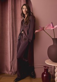 Jedwabny komplet piżamowy w głębokim burgundzie, z długimi rękawami, kieszonką na piersi i elastyczną talią w spodniach. Otoczony różowymi kwiatami i wazonami.