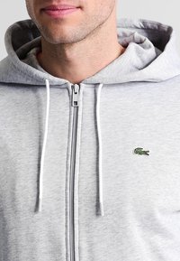 Felpa grigia con zip realizzata in morbido cotone, caratterizzata da un cappuccio con cordino e un piccolo logo di coccodrillo ricamato sul petto.