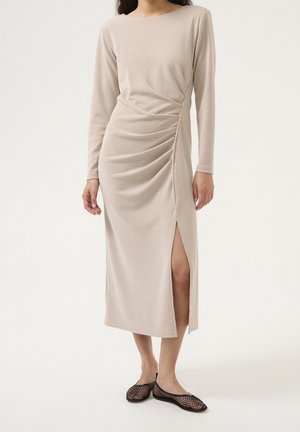 Vrouw draagt een beige midi-jurk met lange mouwen, zijkant rimpelingen en een hoge split, gecombineerd met zwarte platte mesh schoenen.