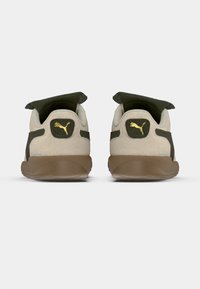 Par de ténis Puma bege e verde-oliva com solas castanhas, vistos de trás, mostrando os logótipos dourados da Puma nas abas do calcanhar.