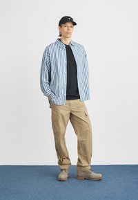 Camisa de botões às riscas azuis e brancas sobre uma t-shirt preta, combinada com calças cargo castanhas claras e botas cinzentas. Modelo usa uma boné preta.