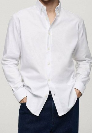 Homme portant une chemise blanche boutonnée, les mains dans les poches d’un pantalon bleu foncé, debout devant un fond clair uni.