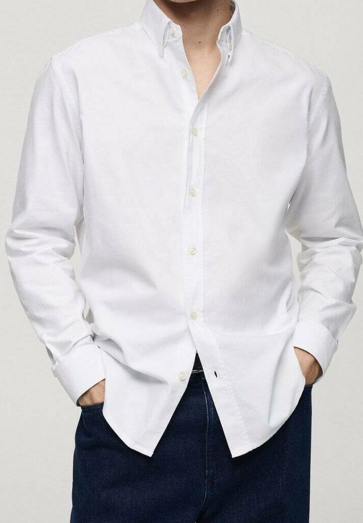 Homme portant une chemise blanche boutonnée, les mains dans les poches d’un pantalon bleu foncé, debout devant un fond clair uni.