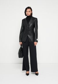 Čierny kožený blejzer s jedným gombíkom, v kombinácii so širokými námorníckomodrými nohavicami. Modelka drží čiernu tašku a nosí čierne topánky s špicatou špičkou.