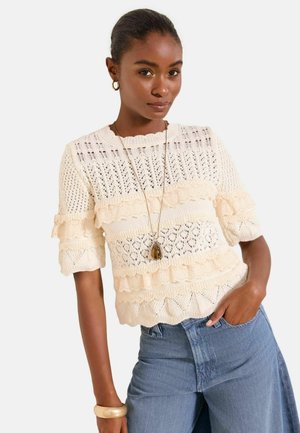 Frau trägt ein cremefarbenes Strickoberteil mit Rüschen, eine lange Anhängerkette, Creolen, ein goldenes Armband und hoch taillierte blaue Jeans, mit einer Hand in der Tasche.