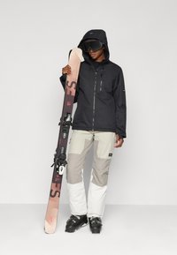Ski suit in black with gray and white pants, holding a brown-sheathed ski. Features a zipper and goggles; snow boots are black with buckles.