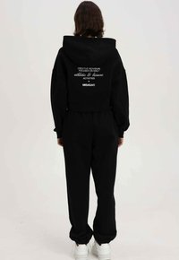 Zwarte cropped hoodie met grote capuchon, geborduurde witte tekst op de achterkant, gecombineerd met losse zwarte broek en witte sneakers.
