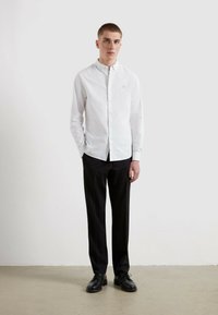 Biała, długosleeve koszula z guzikiem z subtelnym logo na klatce piersiowej, w połączeniu z czarnymi spodniami i czarnymi butami. Prosty design i dopasowany krój.