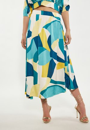Donna che indossa una gonna midi con fantasia astratta colorata, vita elastica e sandali dorati con cinturini e tacco alto, in piedi su sfondo bianco.